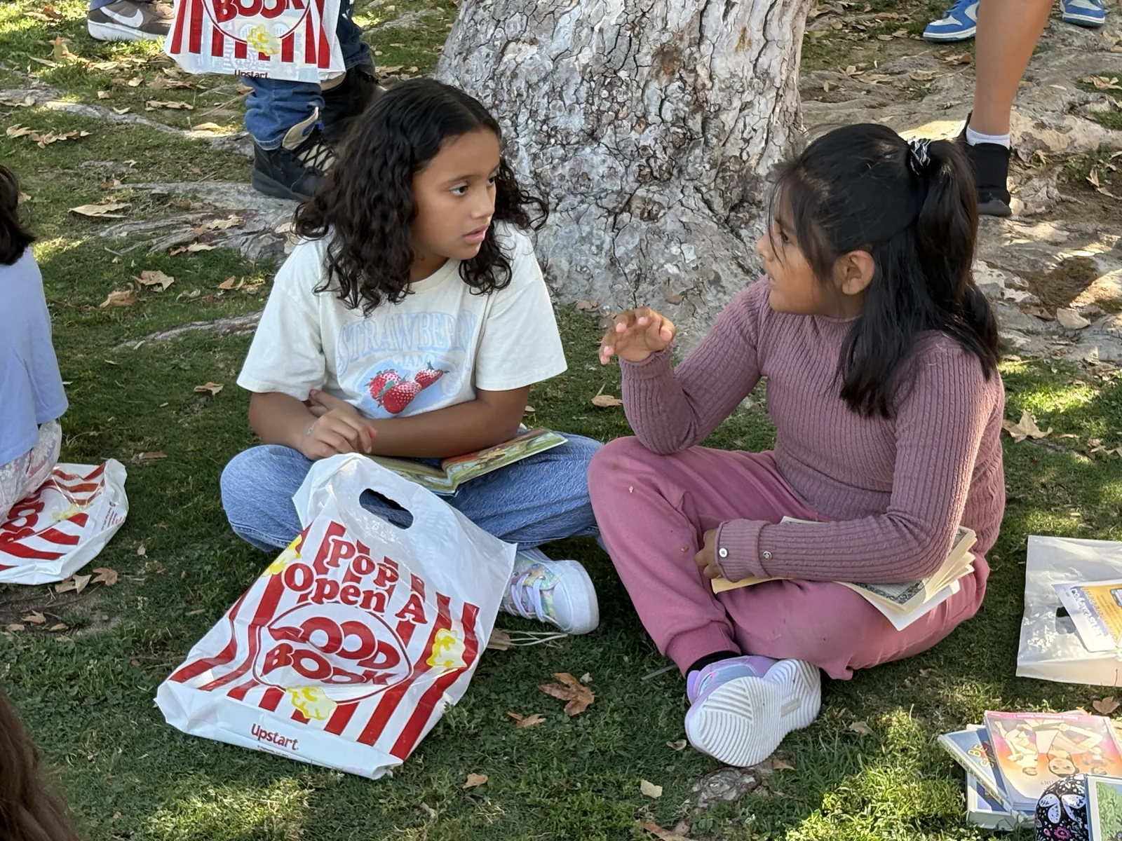 Two students sit on the grass with their books, talking with each other